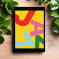 An Apple iPad 7th Gen A2197 10.2" 32GB WiFi Black rests on a wooden surface amid green leaves, its vivid abstract display featuring bold red, pink, blue, and green letters over a yellow background. AU stock, Apple tablet with warranty.