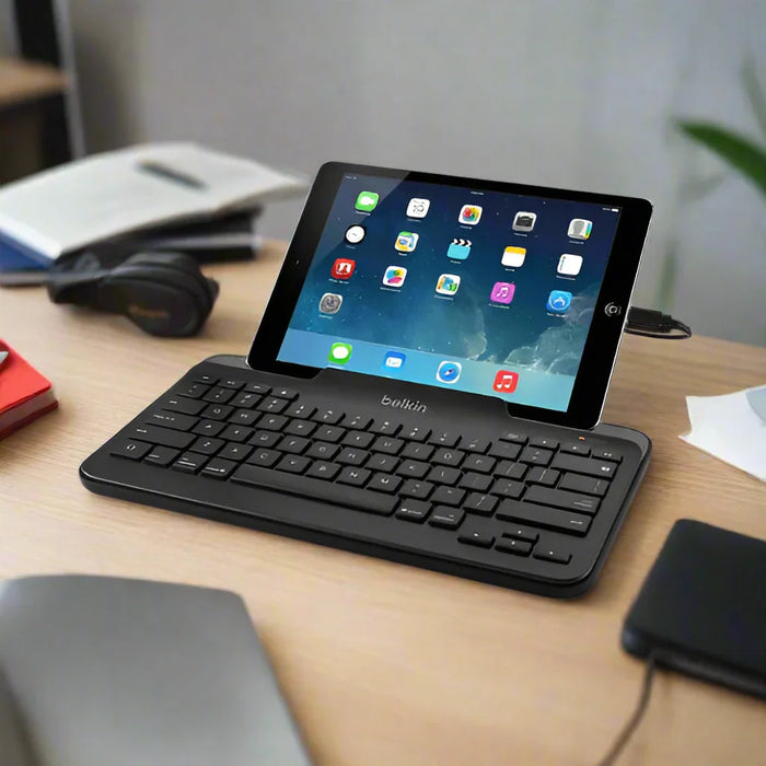 An iPad with app icons is propped on a refurbished Belkin B2B130 Tablet Keyboard with Stand for iPad by Belkin, resting on a wooden desk among books, headphones, and office supplies.