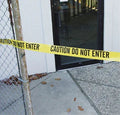 Yellow caution tape reading 'CAUTION DO NOT ENTER' across a glass door entrance near chain-link fence