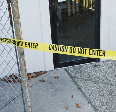 Yellow caution tape reading 'CAUTION DO NOT ENTER' across a glass door entrance near chain-link fence
