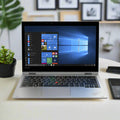 A silver Lenovo ThinkPad Yoga L390 i5 8265U (8GB, 256GB SSD, 14" Touch, W11H) sits on a white desk with the Windows start menu displayed. Nearby are a small potted plant, smartphone, and wallet; leafy decor is visible in the background.